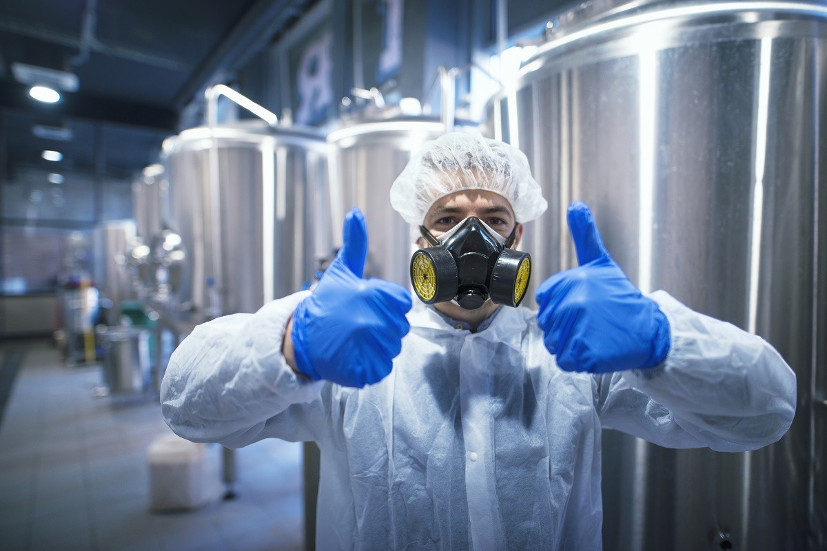 Professional technologist expert in protective white uniform with hairnet, mask and gloves holding both thumbs up.